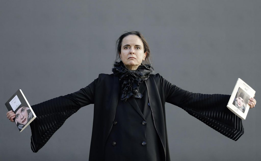 Amelie Nothomb con los brazos extendidos sosteniendo libros, vistiendo abrigo negro sobre fondo gris neutro