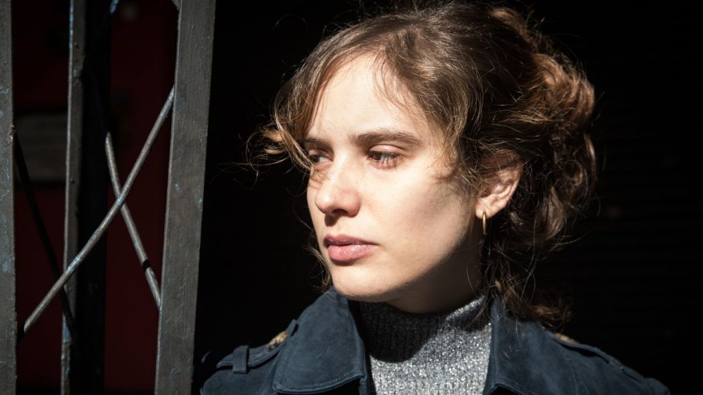 Camila Fabbri de perfil, con cabello recogido y abrigo oscuro, en ambiente teatral con iluminación dramática