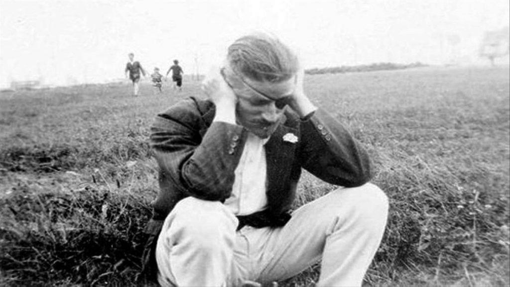 James Joyce en campo abierto, tocándose la cabeza con gesto pensativo, personas al fondo borrosas