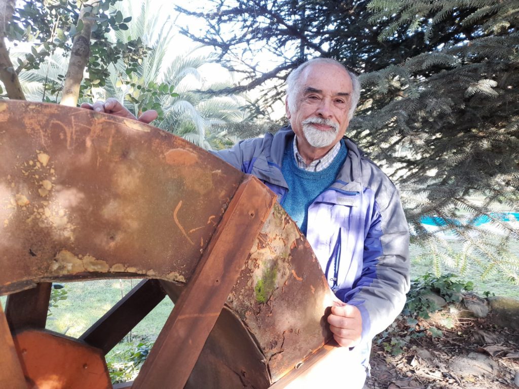Juan Mihovilovich sostiene una pieza de metal oxidado en un jardín con árboles de fondo