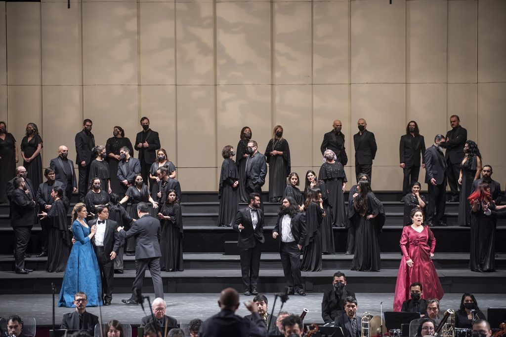 Elenco en escena durante presentación de ópera, con actores en vestuarios oscuros y destacadas figuras en azul y rosa