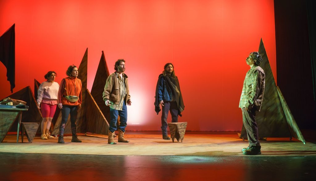 Escena teatral con actores en escenario iluminado de rojo, algunos de pie y otros sentados, con vestuario variado