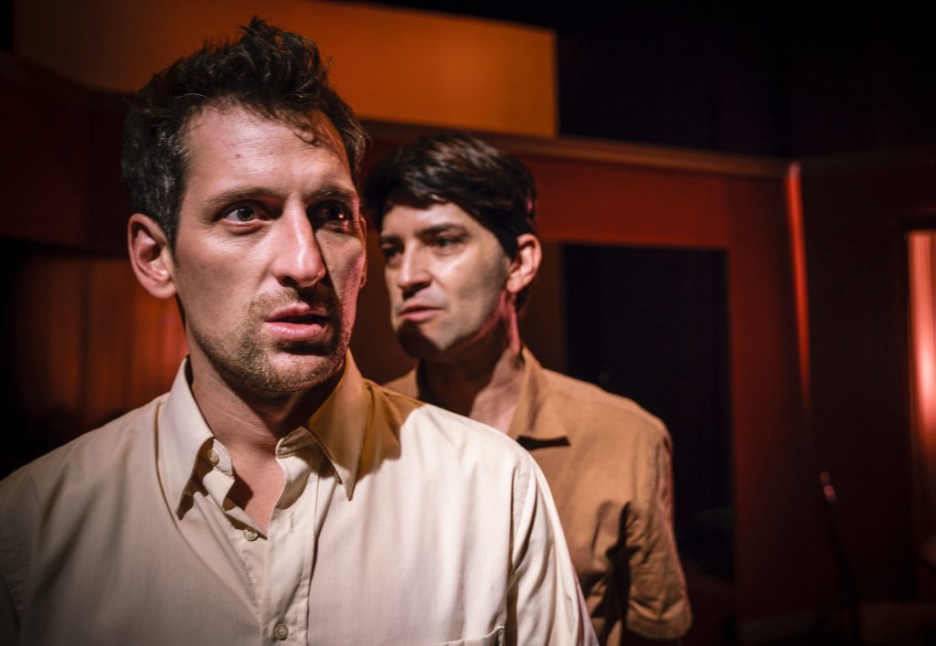 Dos hombres en escena teatral con iluminación cálida naranja. El del frente viste camisa clara, expresión seria. Fondo