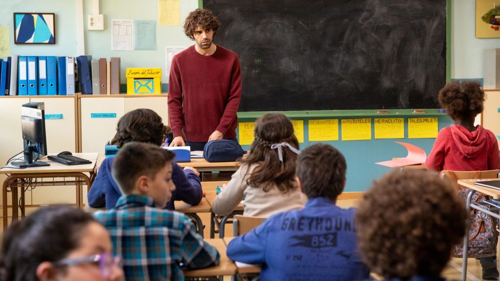 Docente en aula roja dirigiéndose a estudiantes de primaria sentados en círculo alrededor de una mesa de trabajo