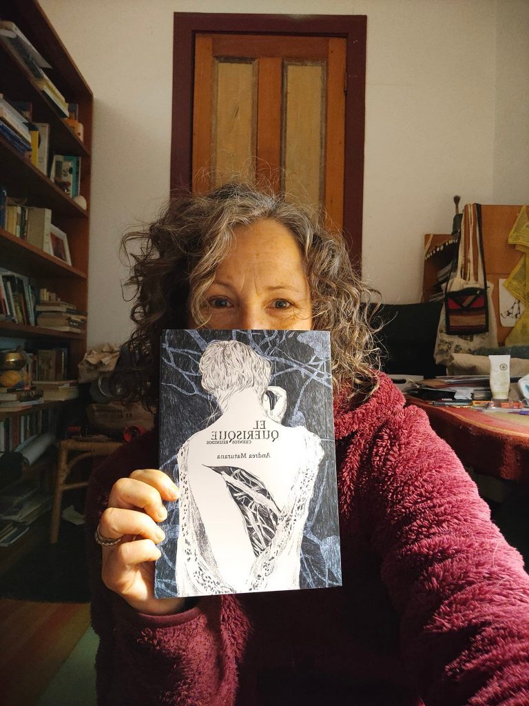 Andrea Maturana sostiene un libro con ilustración de rostro, en su oficina con estanterías de fondo