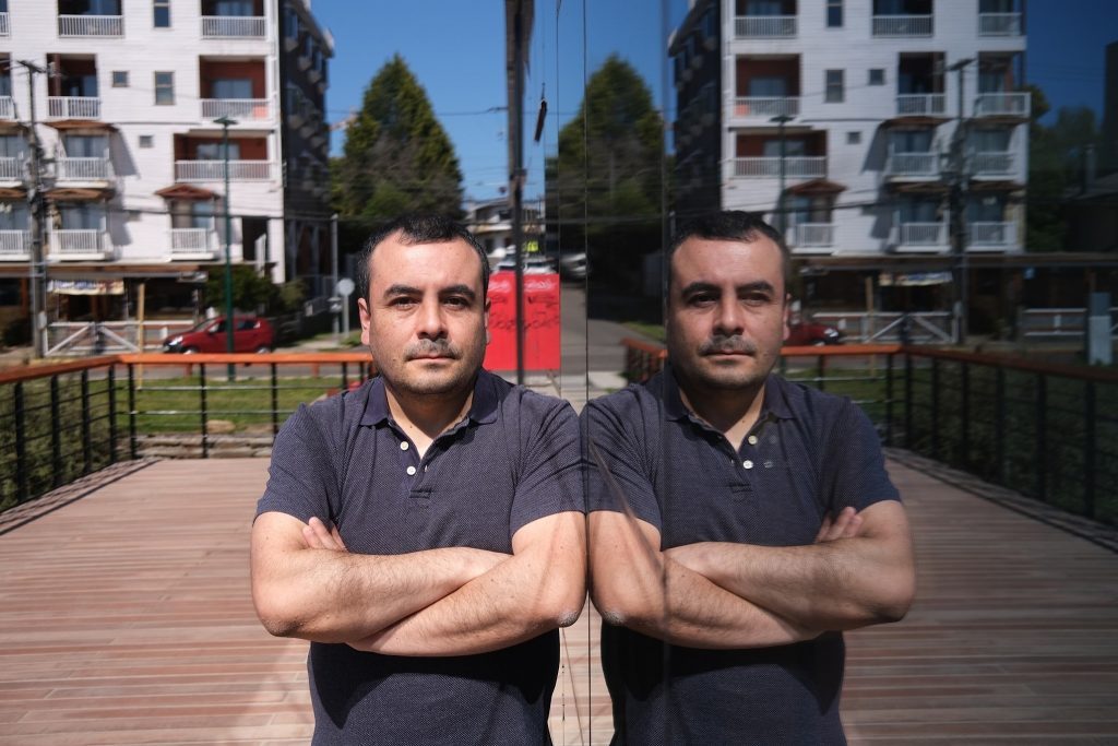 Dos hombres jóvenes con brazos cruzados en una terraza de madera, edificios residenciales al fondo