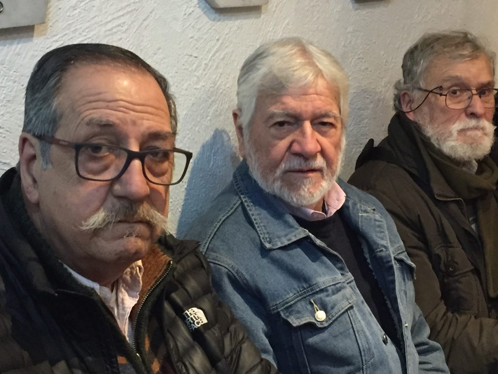 Tres hombres de mediana edad en interior, el del centro viste camisa de mezclilla, expresión seria y reflexiva