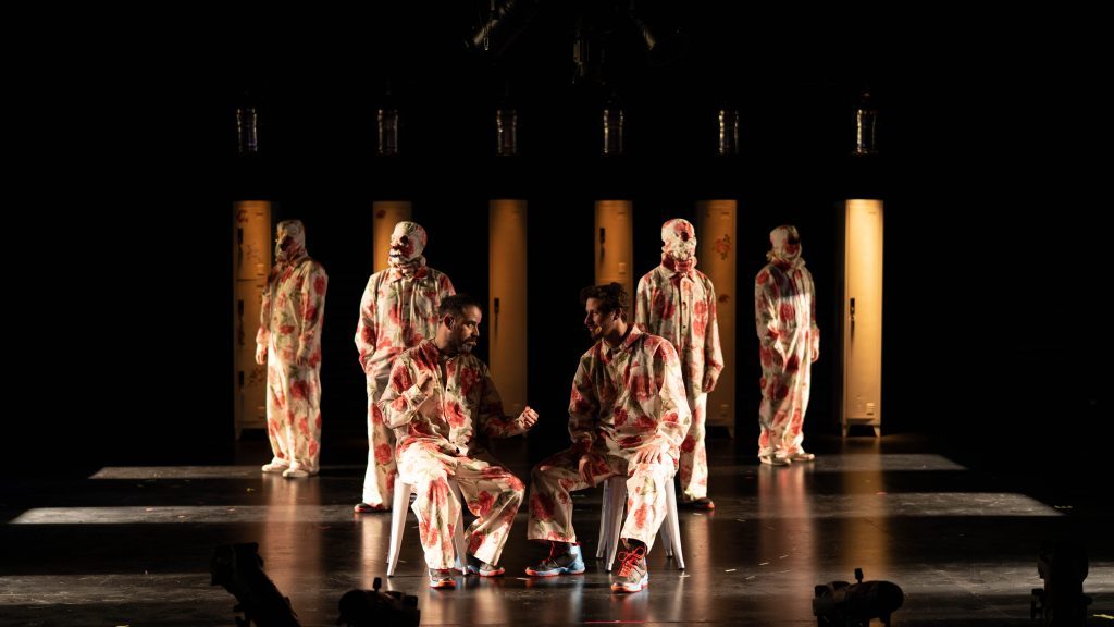 Escena teatral con actores en trajes de época bajo luces doradas, posados en formación sobre escenario oscuro