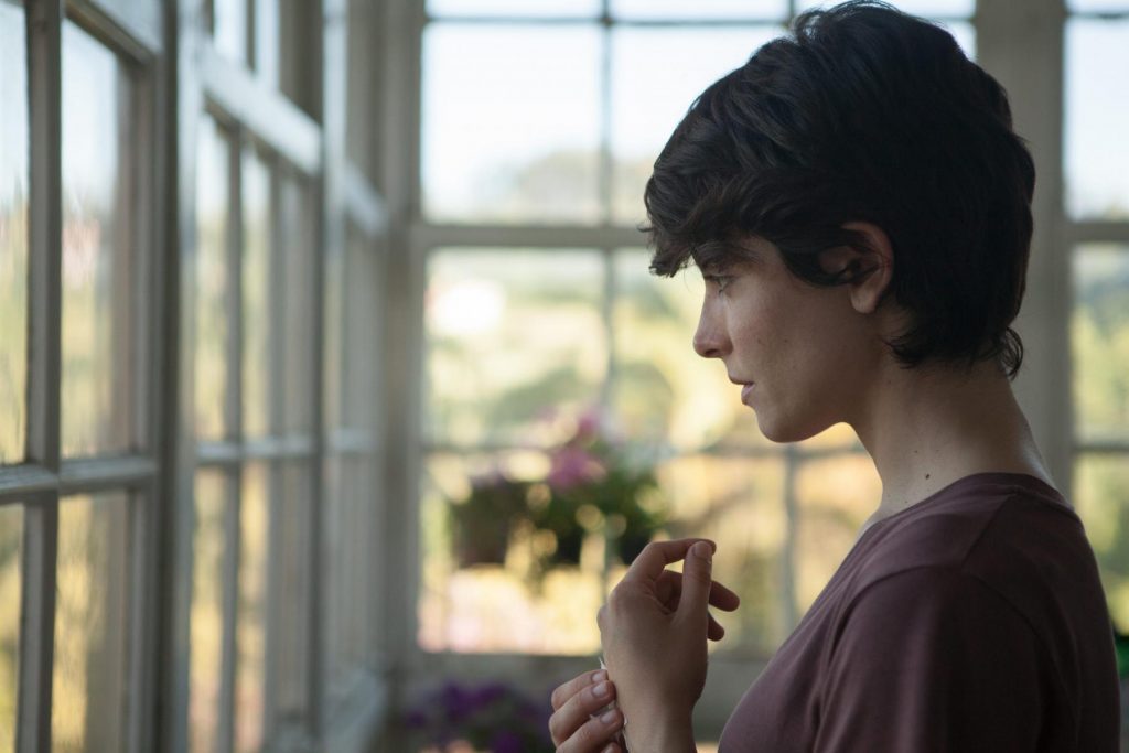 Joven mujer de perfil junto a ventana con marcos de madera, mirando hacia afuera con expresión contemplativa