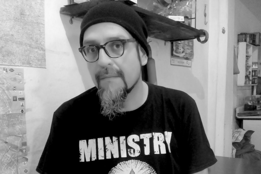 Patricio Jara, con gafas y gorro negro, viste camiseta con logo de Ministry en estudio con documentos de fondo