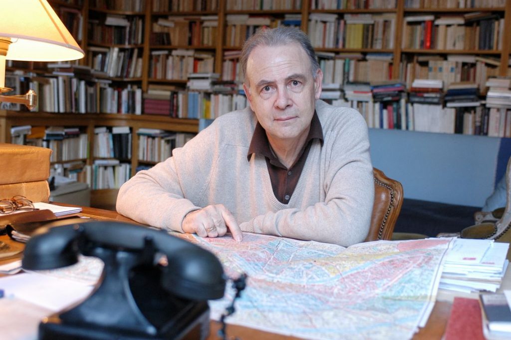 Patrick Modiano en su estudio con librerías al fondo, trabajando sobre mapas y documentos sobre su escritorio