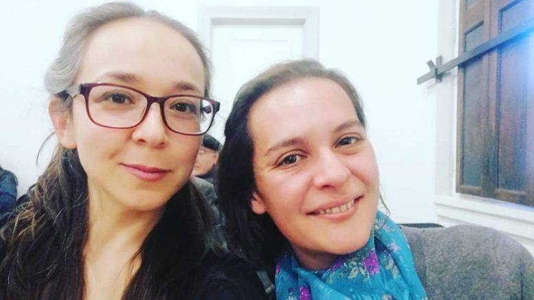 Dos mujeres sonriendo a la cámara en interiores, una con gafas y cabello largo oscuro