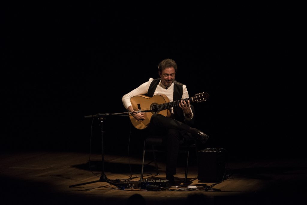 Guitarrista flamenco tocando en escena sobre fondo negro, con iluminación de spot, durante presentación en vivo