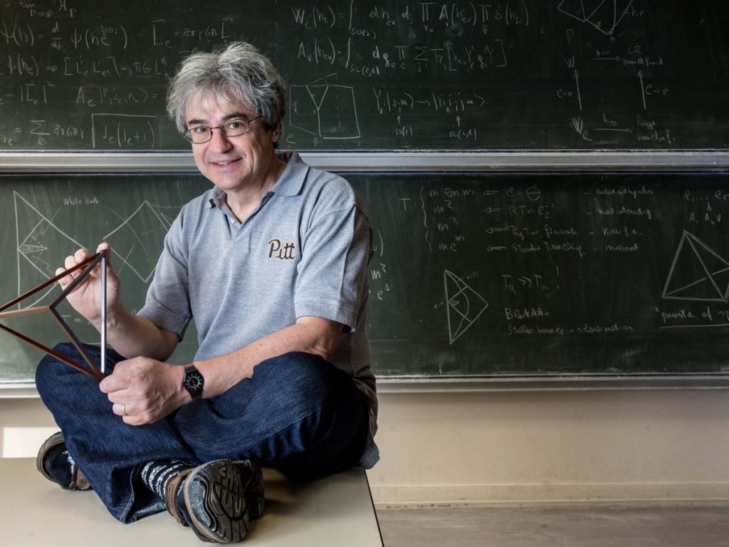 Carlo Rovelli sentado frente a pizarra con ecuaciones, sosteniendo libro, sonriendo hacia cámara en aula