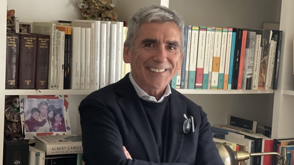Carlos Peña sonriendo frente a estantería con libros, vistiendo camisa negra en interior de biblioteca