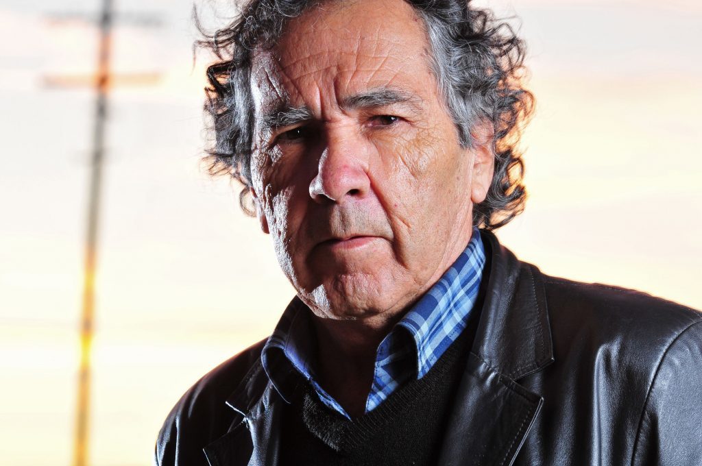 Hernán Rivera Letelier, escritor chileno de cabello gris ondulado, viste chaqueta negra sobre camisa azul en exterior