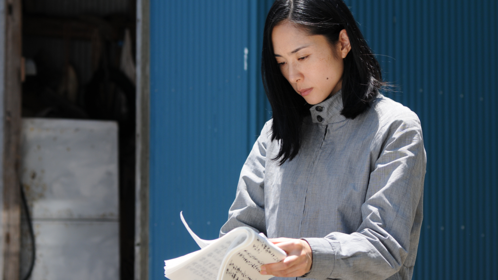 Mujer sostiene un libro abierto mientras lee, con puerta azul de fondo, en escena de reflexión literaria