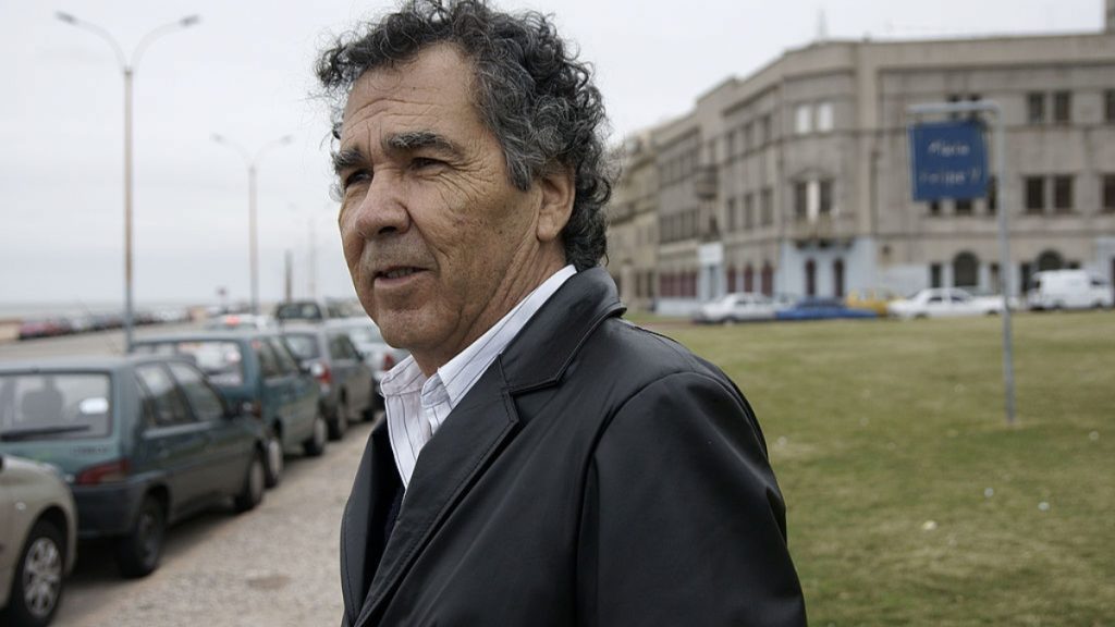 Hernán Rivera Letelier, escritor chileno, en traje oscuro frente a edificio y estacionamiento