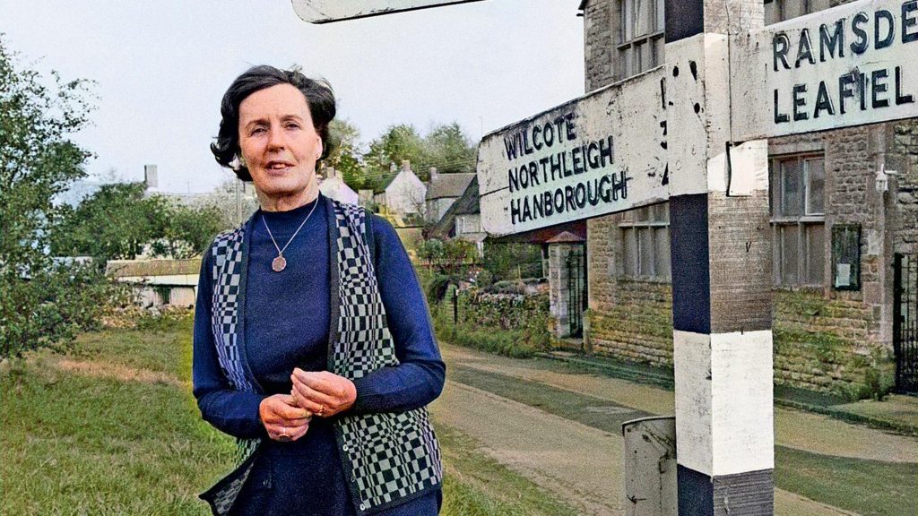 Escritora Barbara Pym frente a señal de estación de tren británica, sonriendo hacia la cámara en jardín