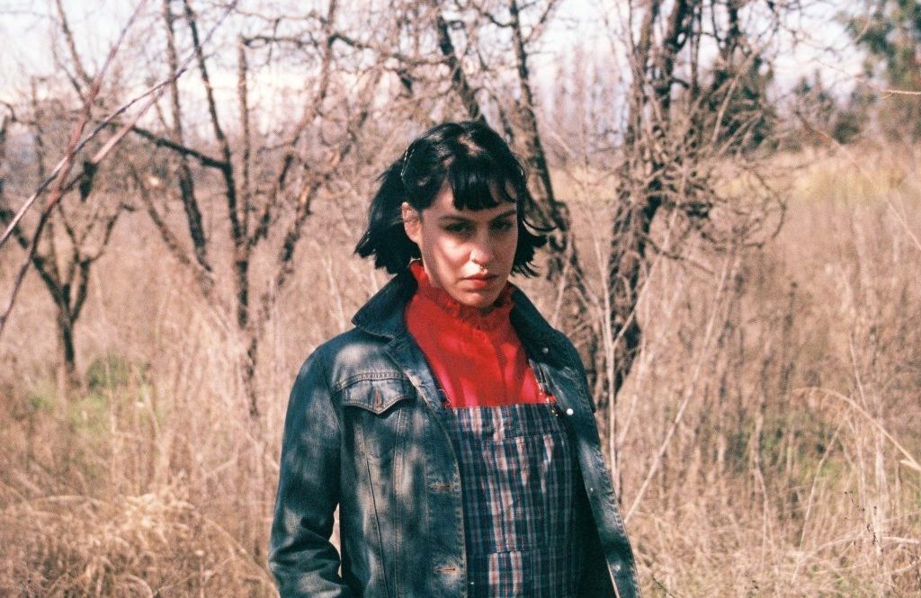 Josefina González de pie en un terreno árido con vegetación seca, vistiendo camisa de cuadros y suéter rojo oscuro