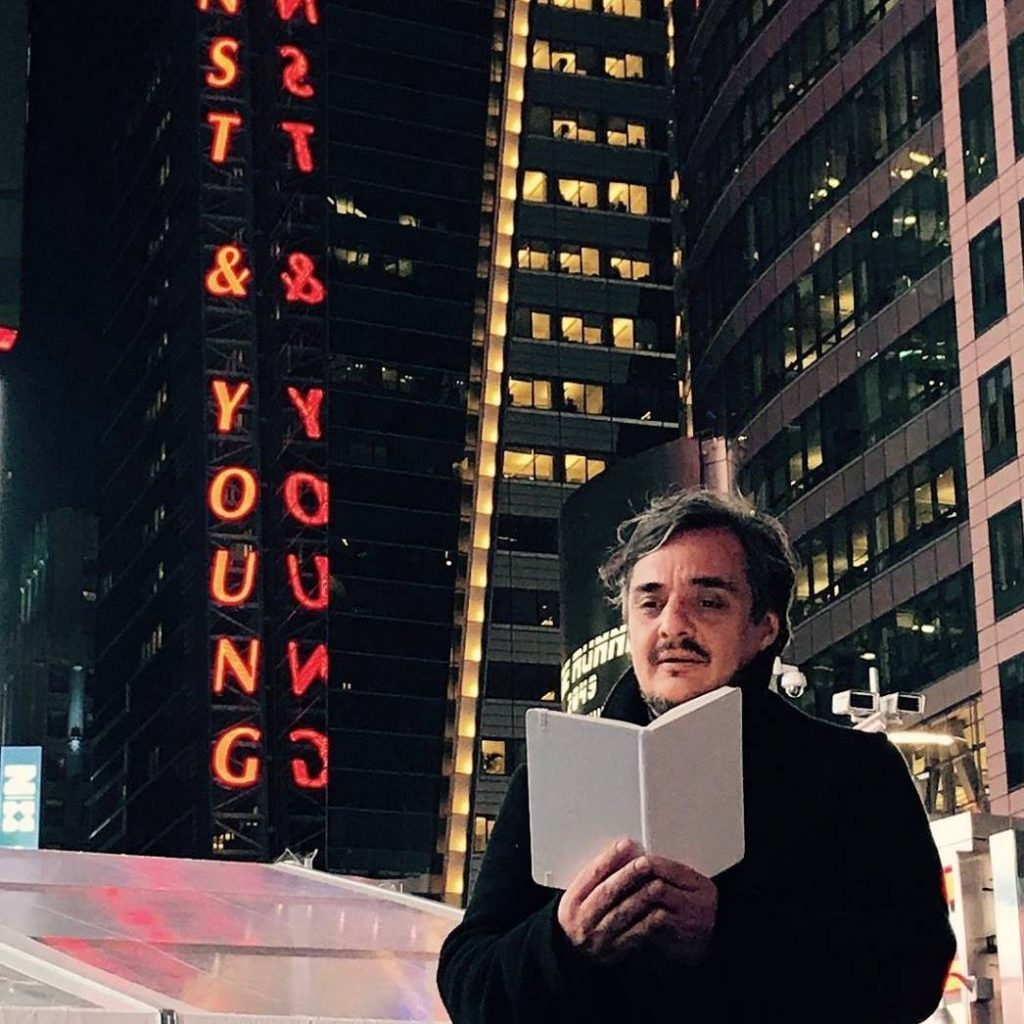 Juan Pablo Meneses sostiene un libro abierto bajo letreros de neón rojo en noche urbana con rascacielos iluminados