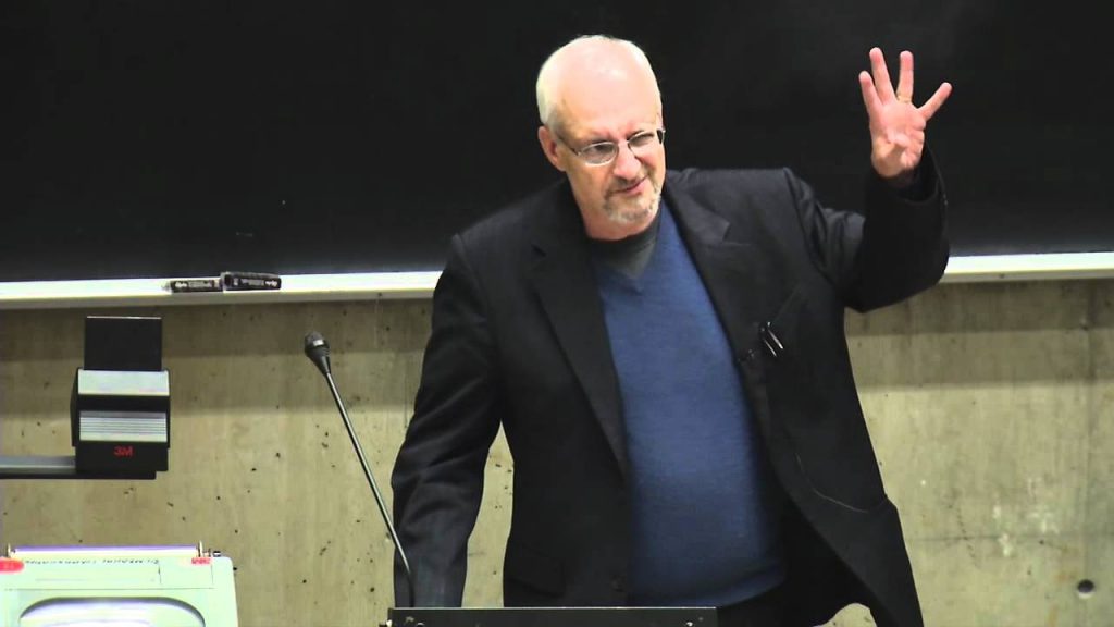 Morris Berman, hombre mayor con gafas, dicta conferencia en aula universitaria frente a pizarra blanca, gesticulando