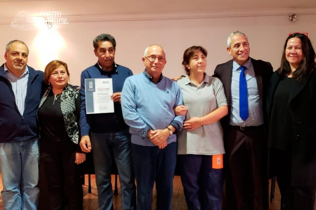 Grupo de ocho personas en interior posando para foto, uno sostiene diploma o certificado, ambiente formal