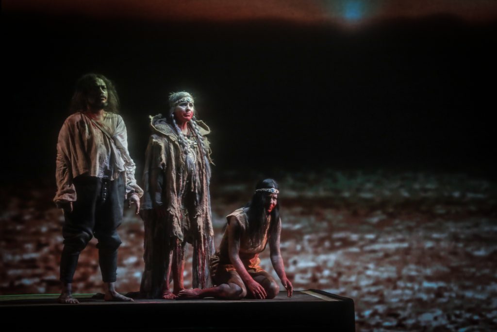 Tres actores en escena sobre tierra árida bajo luz dramática, con vestuario de época en tonos ocres y marrones