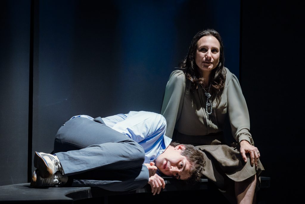 Dos actores en escena teatral: mujer sentada de perfil con abrigo marrón, hombre acostado en el piso con camisa azul