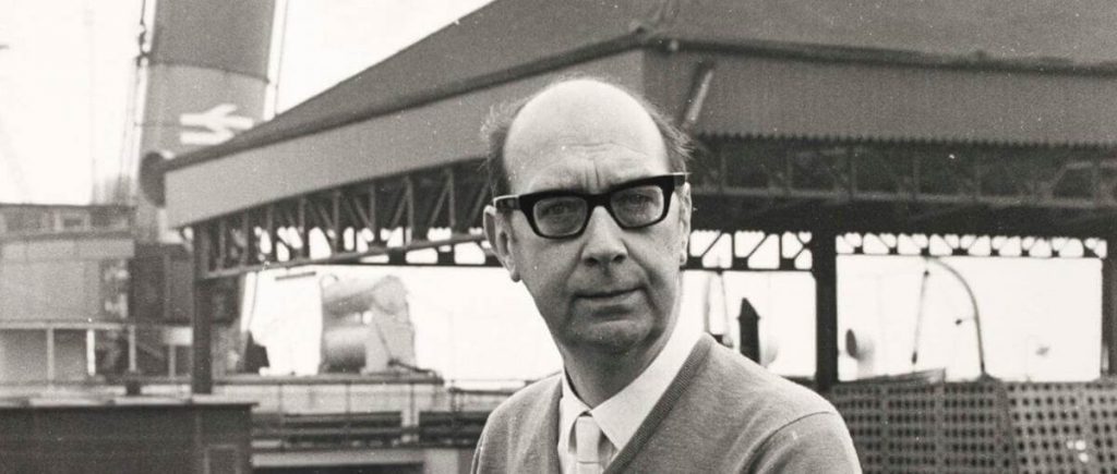 Philip Larkin, poeta británico, de perfil tres cuartos con gafas oscuras y traje claro frente a estructura industrial