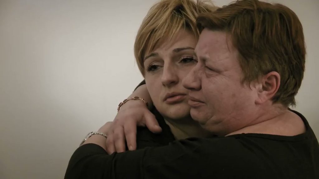 Dos personas en abrazo emotivo, con expresiones de dolor y vulnerabilidad, sobre fondo neutro