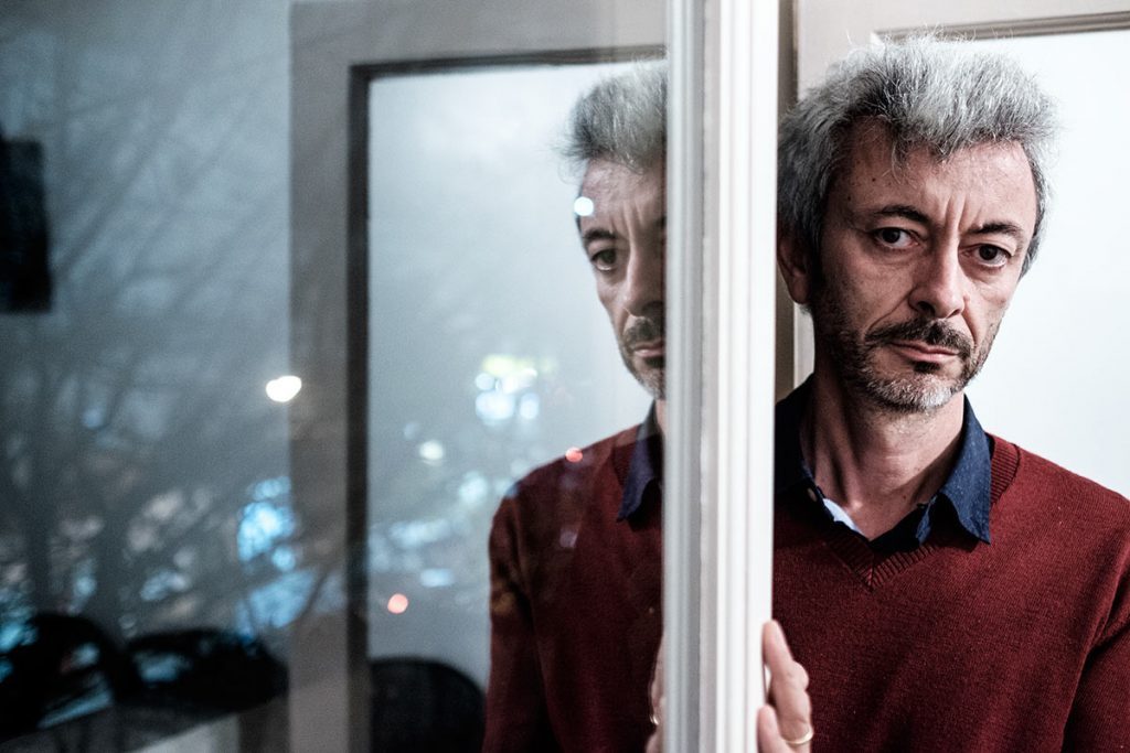 Hombre de cabello gris y barba, vistiendo suéter burdeos, mirando hacia la ventana con expresión reflexiva en ambiente