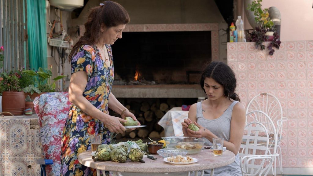 Dos mujeres en una terraza mediterránea; una sirve comida a la otra sentada a la mesa con plantas de fondo