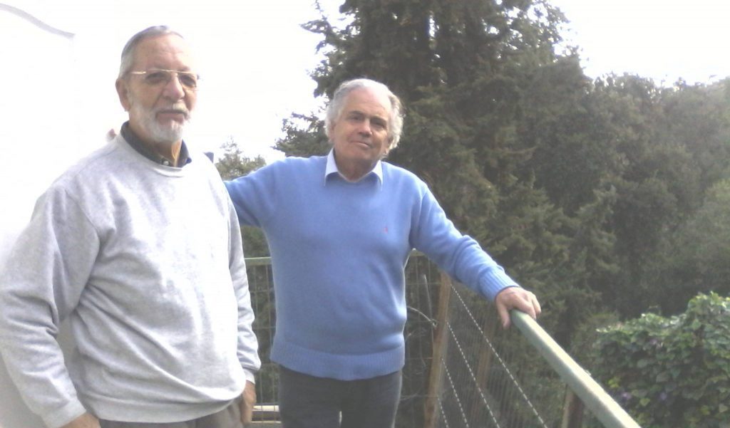 Dos hombres en una terraza con barandilla de madera, rodeados de vegetación frondosa al fondo