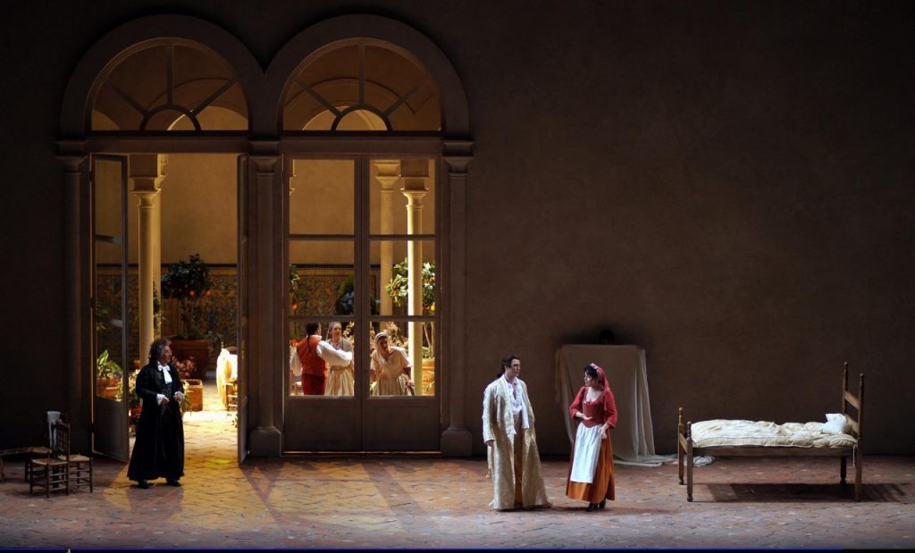Escena teatral nocturna con fachada iluminada, personajes en primer plano y decorados de época en un escenario