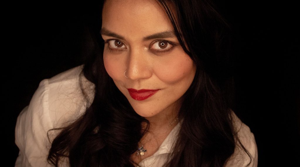 Retrato de Laura Baeza, escritora, con cabello largo castaño oscuro, maquillaje definido y blusa clara sobre fondo negro