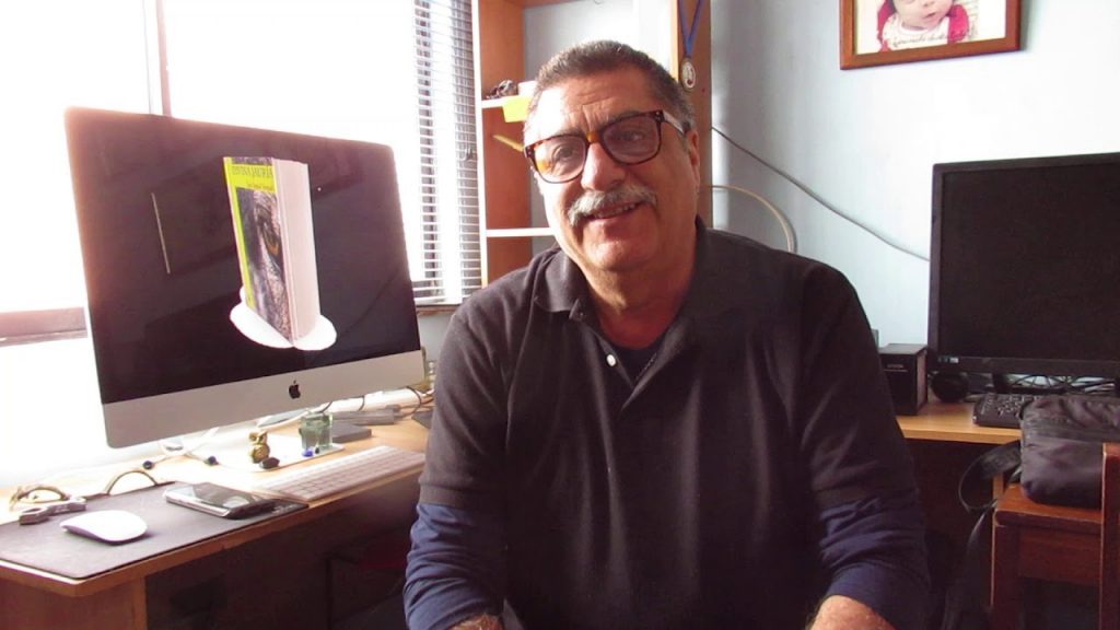 Luis Seguel en su oficina, frente a computadora, con gafas y suéter oscuro, rodeado de materiales de cine y literatura