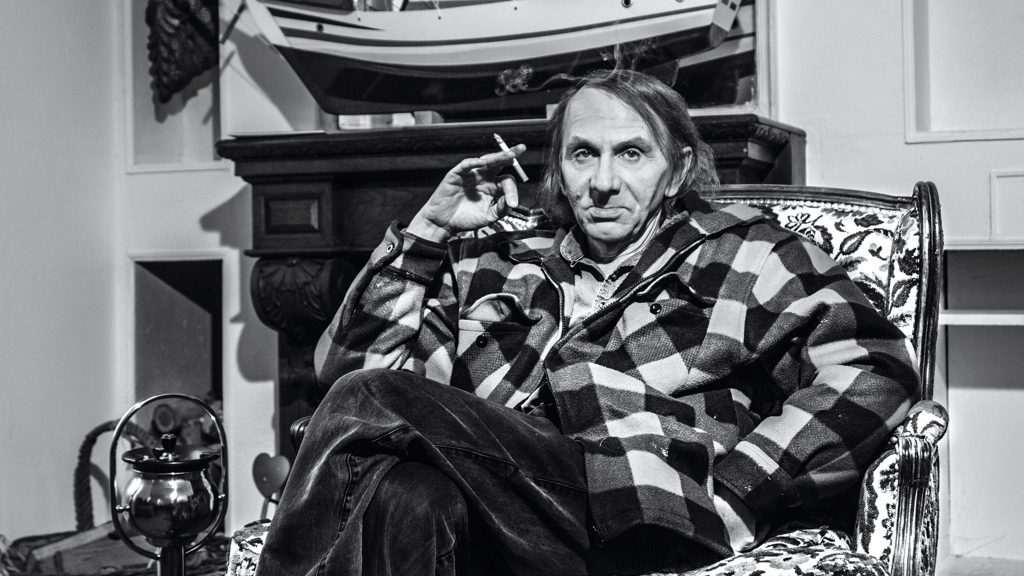 Michel Houellebecq recostado en una silla, vistiendo camisa a cuadros, en blanco y negro