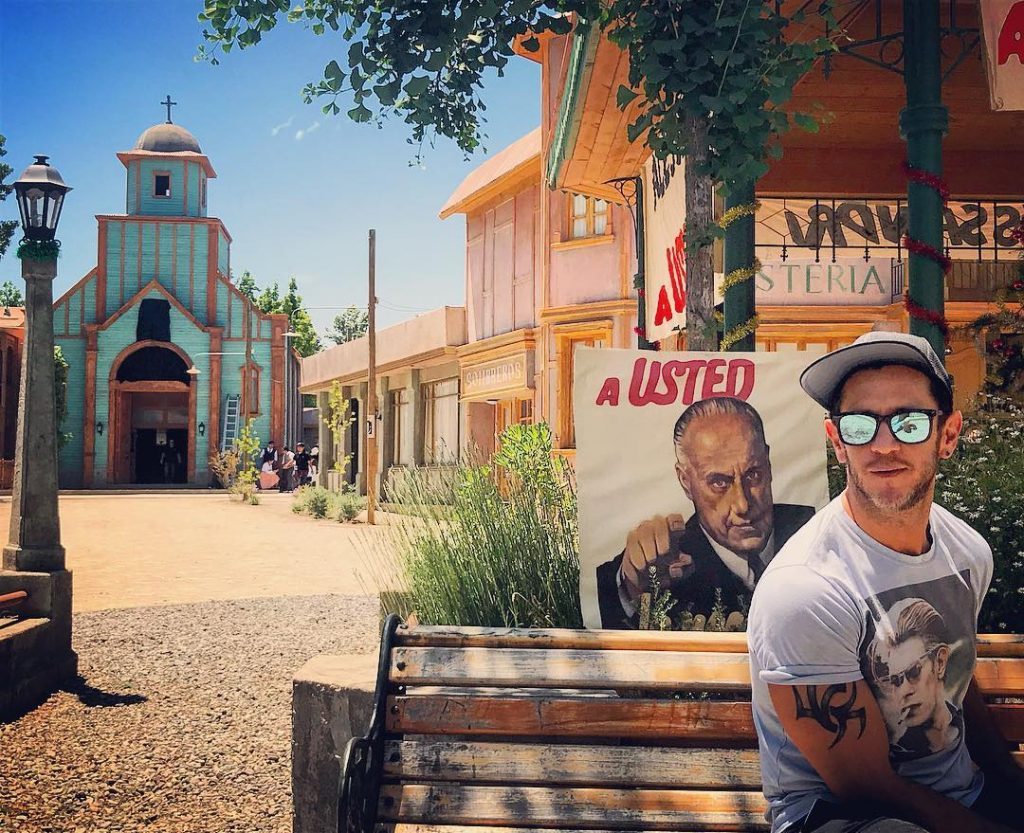 Hombre con lentes sonríe frente a cartel de película en set de cine del oeste, pueblo antiguo de fondo