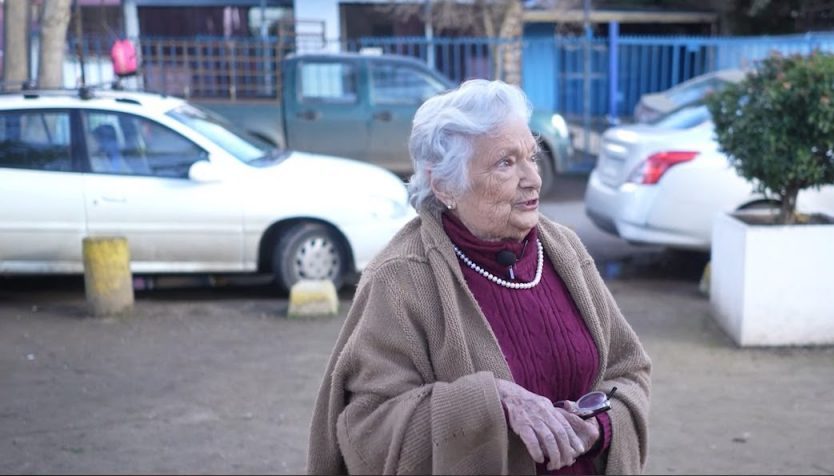 Patricia Schaffner, mujer mayor de cabello blanco, viste abrigo marrón y suéter granate en estacionamiento