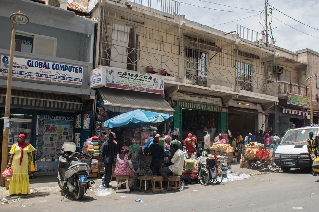 Calle concurrida de Dakar con comercios, vendedores ambulantes, motocicletas y peatones bajo sombrillas en clima