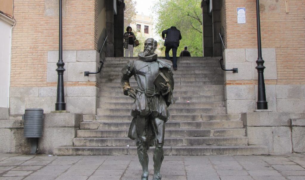 Estatua de Miguel de Cervantes en Toledo, figura de bronce en escalinata de piedra entre edificios históricos