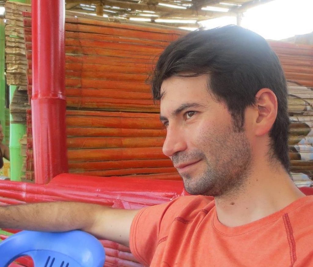 Hombre de cabello oscuro sonriendo dentro de un espacio con paredes de madera roja y estructura metálica rosa