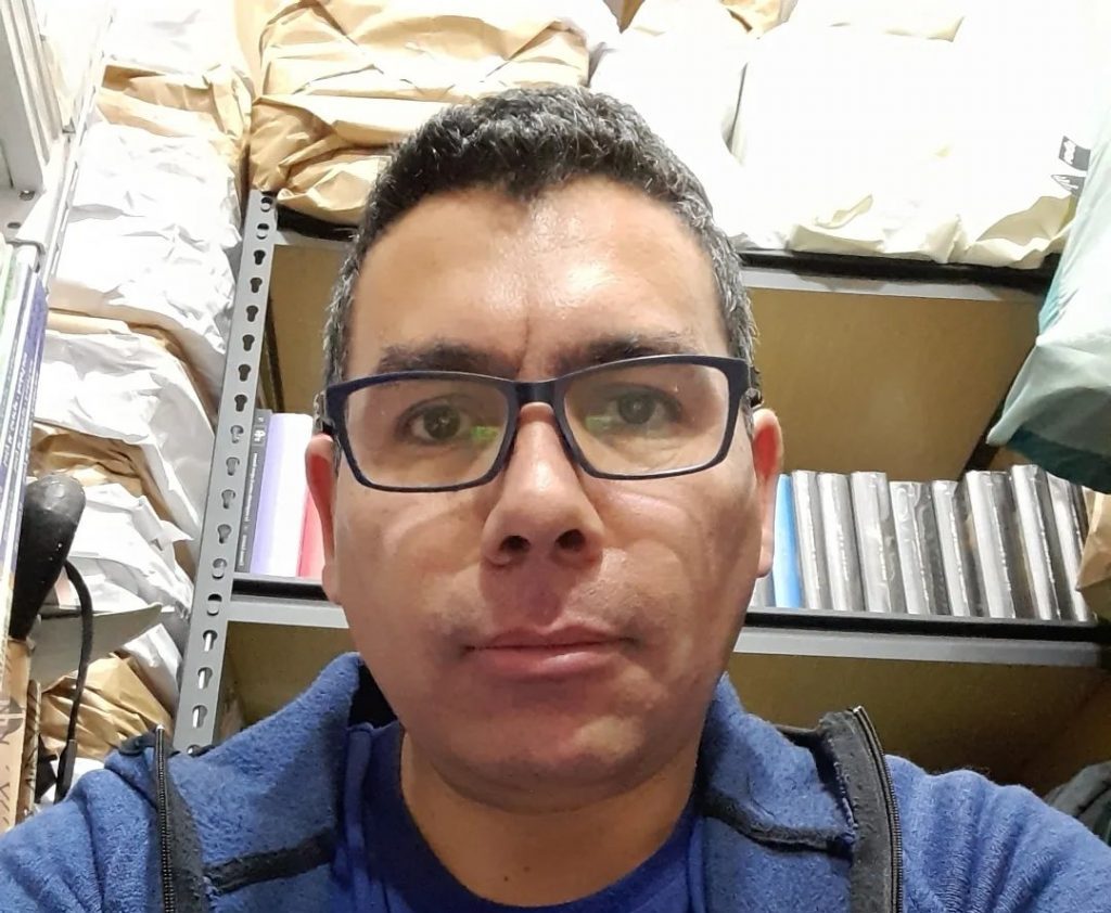 Hombre de lentes oscuros y camisa azul en interior con estanterías de libros al fondo