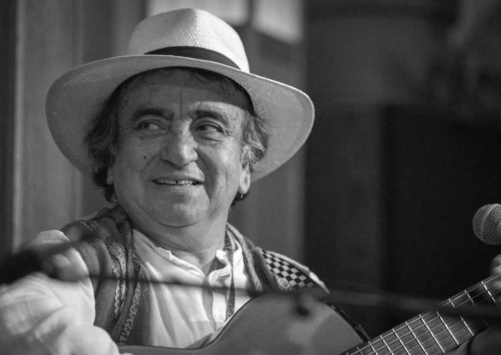 Hombre con sombrero de ala ancha y camisa a cuadros sosteniendo una guitarra, sonriendo hacia la cámara en fotografía