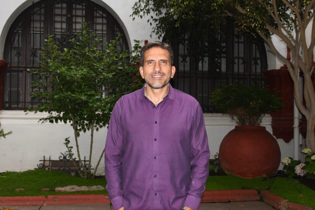 Cristián Brito Villalobos en jardín con arquitectura colonial, vistiendo camisa morada