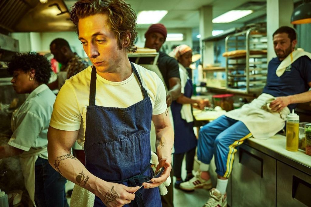 Joven cocinero con uniforme azul prepara comida en cocina profesional abarrotada mientras otros trabajan al fondo