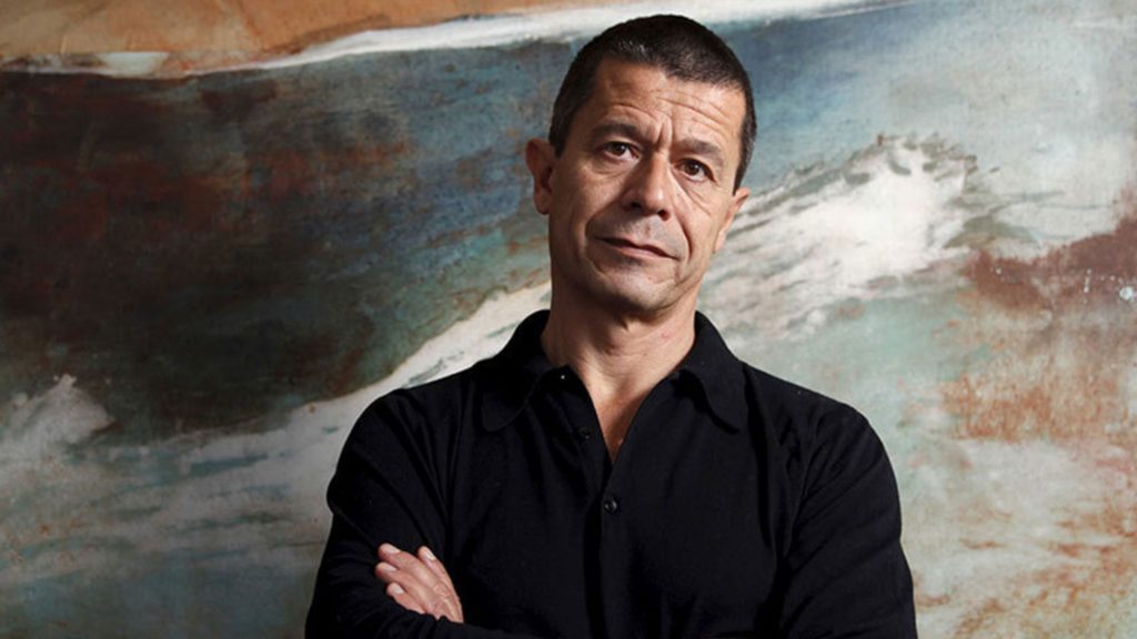 Emmanuel Carrère, escritor francés, de brazos cruzados frente a un cuadro abstracto con tonos ocres y azules