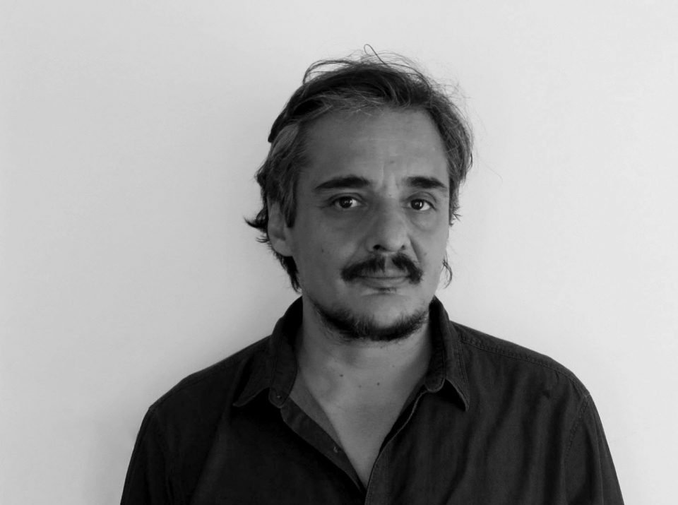 Retrato en blanco y negro de Juan Pablo Meneses, escritor chileno, con bigote prominente y camisa oscura sobre fondo