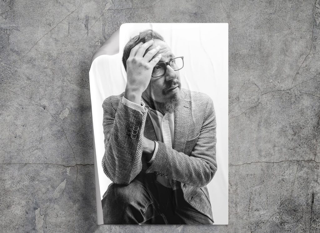 Víctor Ilich sostiene una cámara fotográfica frente a su rostro, retrato en blanco y negro sobre fondo gris texturado